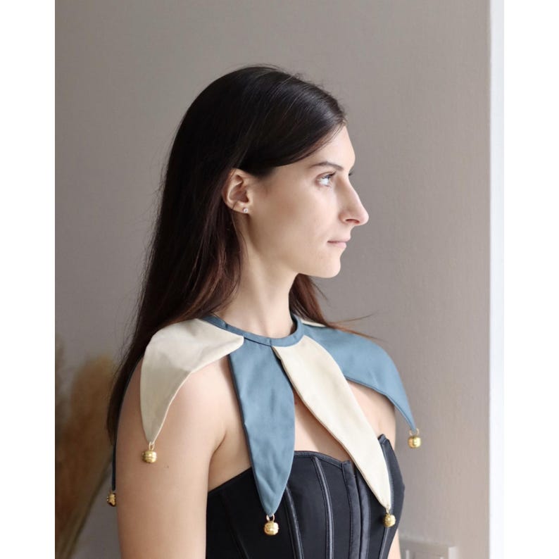 May include: A blue, cream, and gold jester collar with bell accents. The collar is made of fabric and features a pointed design. The jester collar is worn over a black corset.