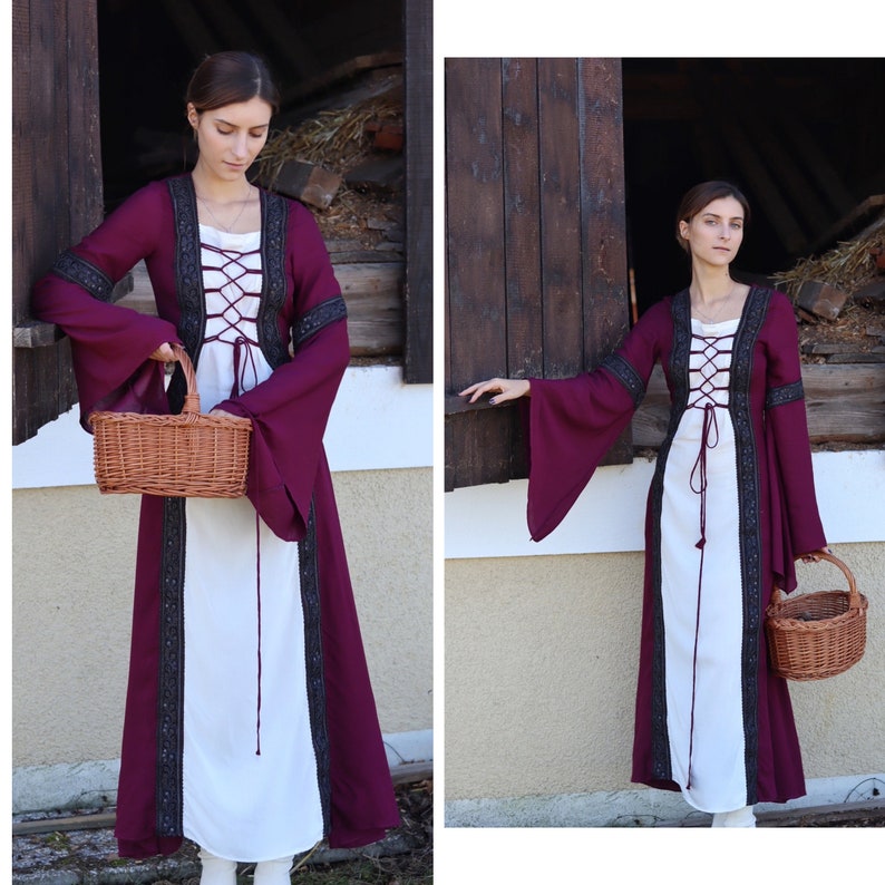 May include: A woman wearing a long, white dress with a burgundy overlay and black lace trim. The dress has a lace-up bodice and long sleeves. She is holding a wicker basket.