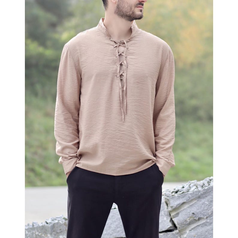 May include: A long-sleeved, beige shirt with a mandarin collar and a lace-up front. The shirt has a textured appearance and is paired with black pants. The overall style suggests a casual, yet refined aesthetic.