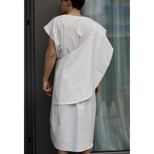 May include: A white, sleeveless tunic and draped garment. The outfit appears to be made of a lightweight fabric. The model is wearing a white tunic and a draped piece of fabric over the shoulder.