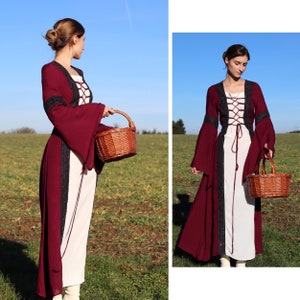 May include: A woman wearing a long, burgundy dress with a white lace bodice and black trim. She is holding a brown wicker basket in her right hand. The dress has long sleeves with bell bottoms. The woman is standing in a field with green grass and a blue sky in the background.