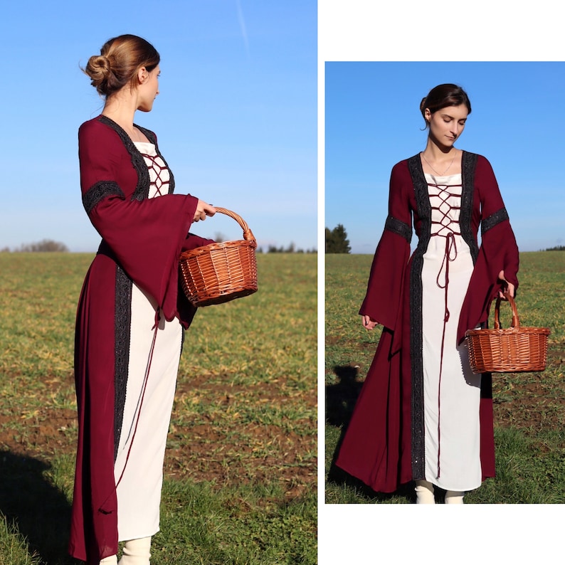 May include: A woman wearing a long, burgundy dress with a white lace bodice and black trim. She is holding a brown wicker basket in her right hand. The dress has long sleeves with bell bottoms. The woman is standing in a field with green grass and a blue sky in the background.