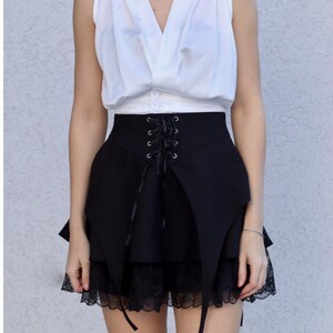 May include: Black skirt with a high waist and lace trim. The skirt features a lace-up detail in the front and a unique asymmetrical design. The skirt is paired with a white sleeveless top.