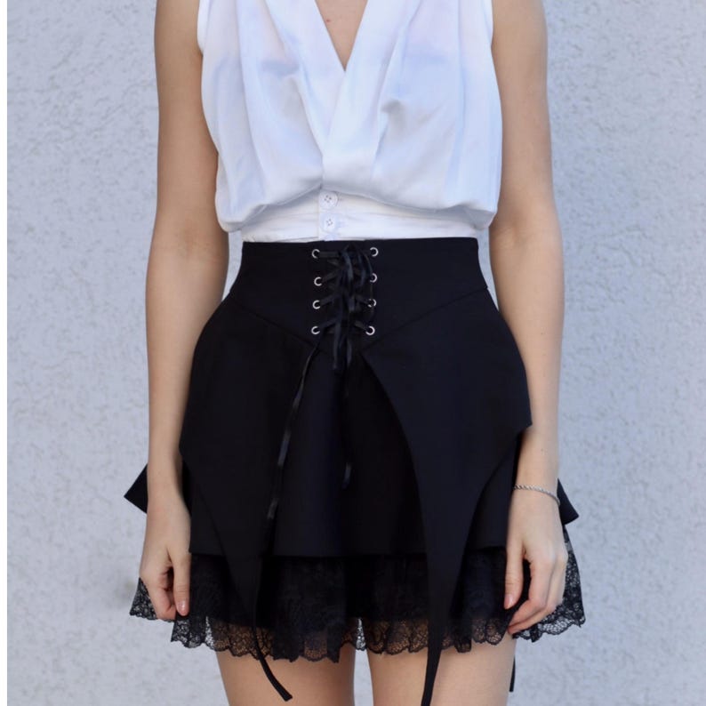 May include: Black skirt with a high waist and lace trim. The skirt features a lace-up detail in the front and a unique asymmetrical design. The skirt is paired with a white sleeveless top.