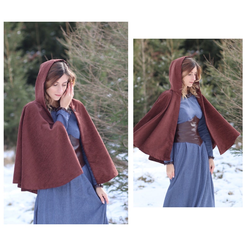 May include: A woman wearing a brown hooded cape over a blue dress. The cape has a long, flowing design and is made of a soft, textured fabric. The woman is standing in a snowy forest setting.