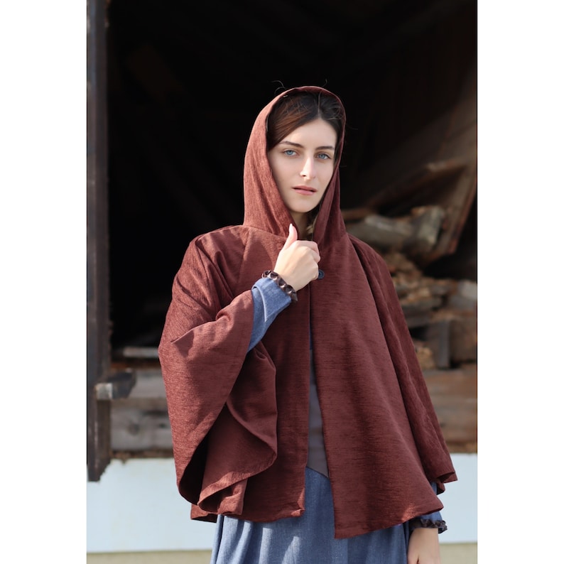 May include: A brown hooded cape with a textured fabric. The cape is worn by a person with long brown hair and blue eyes.