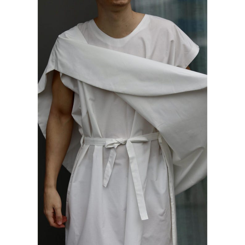 May include: A white cotton tunic with a matching belt tied at the waist. The tunic has short sleeves and a draped shoulder piece. The fabric appears lightweight and flowing, suggesting a casual or artistic style.