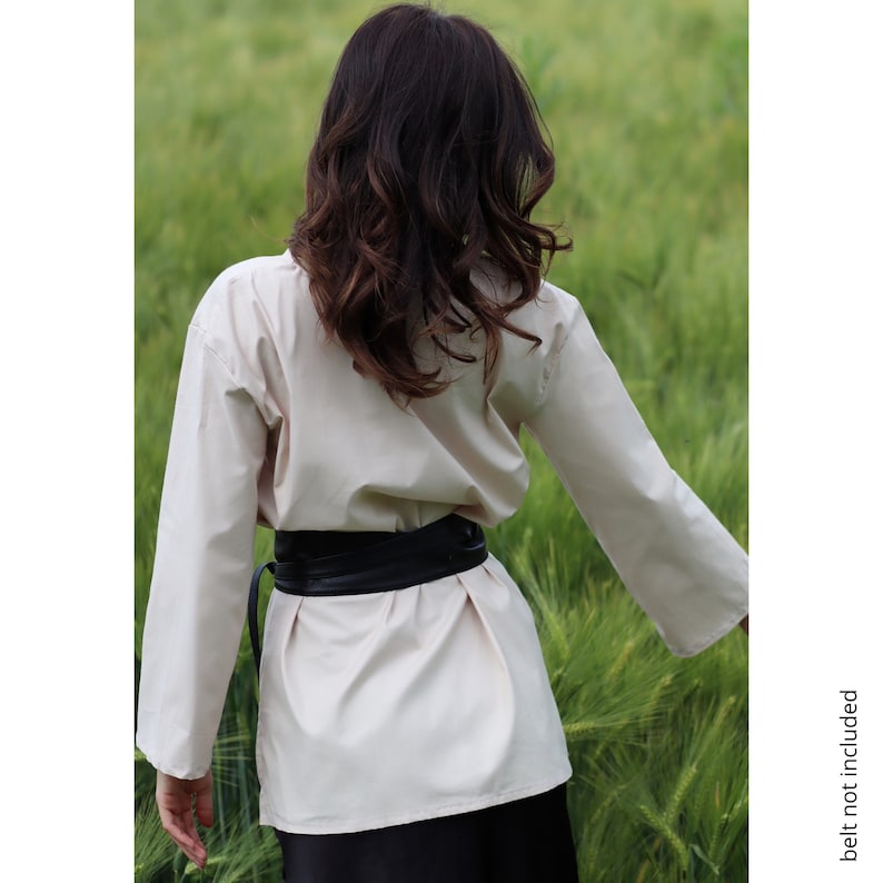 May include: A cream-colored shirt with long sleeves and a black belt tied around the waist. The shirt is worn by a person with long brown hair standing in a field of tall green grass.