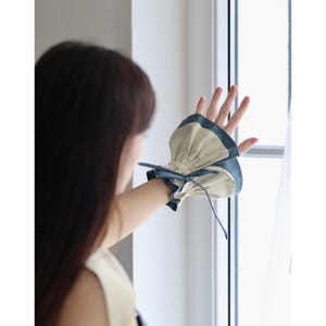 May include: A beige and teal wrist cuff with a gathered design and a teal bow. The cuff is worn on the wrist and has a contrasting teal trim. The cuff is made of a soft fabric.