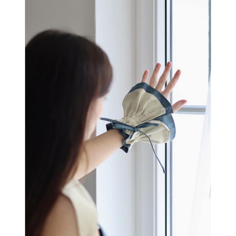 May include: A beige and teal wrist cuff with a gathered design and a teal bow. The cuff is worn on the wrist and has a contrasting teal trim. The cuff is made of a soft fabric.