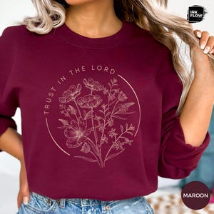 May include: Maroon sweatshirt with a floral design inside a circle and the text "TRUST IN THE LORD". The sweatshirt has long sleeves and a crew neck. The word "MAROON" is printed on the bottom right corner.