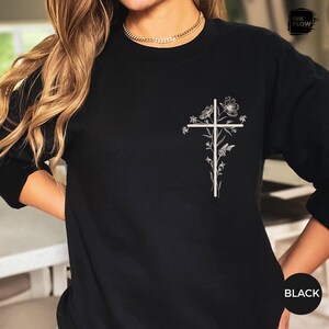 May include: Black long-sleeved sweatshirt with a white floral cross design on the front. The cross is decorated with flowers and butterflies. The word "BLACK" is printed on the bottom right.