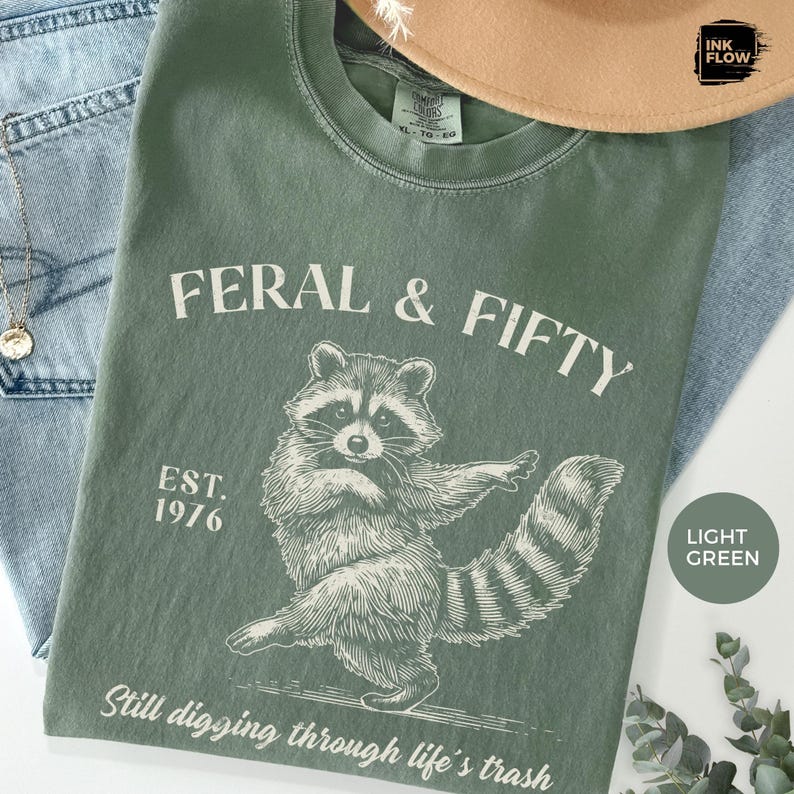 May include: Light green t-shirt with the text "FERAL & FIFTY" and a raccoon graphic. The shirt also features the text "EST. 1976" and "Still digging through life's trash". A tan hat and blue jeans are in the background.