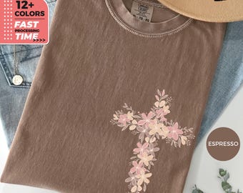 May include: Espresso-colored t-shirt with a floral cross design. The cross is made of pink and peach-colored flowers and leaves. The shirt is folded on top of blue jeans. The image also includes text that says "12+ COLORS" and "FAST PROCESSING TIME".