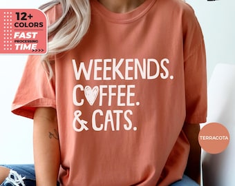 May include: A terracotta colored t-shirt with the text "Weekends. Coffee. & Cats." in white. The text includes a small heart symbol in the word "Coffee".
