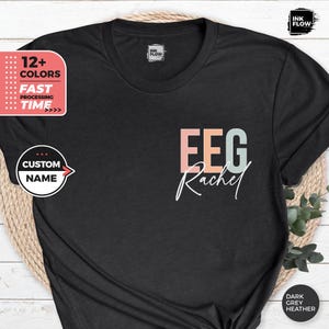 Op de afbeelding: Een zwart T-shirt met de tekst "EEG Rachel" in kleurrijke letters. De tekst is in een moderne lettertype stijl.