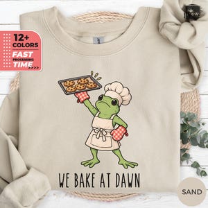 Könnte beinhalten: Ein sandfarbenes Sweatshirt mit Rundhalsausschnitt und einer Grafik eines grünen Frosches mit weißer Kochmütze und Schürze. Der Frosch hält ein Backblech mit vier Schokoladenkeksen und trägt einen rot-weiß karierten Ofenhandschuh. Text: WE BAKE AT DAWN.