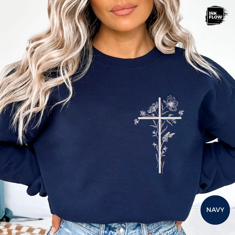 May include: Navy blue sweatshirt with a white floral cross design. The cross is decorated with flowers and leaves. The sweatshirt has a crew neck and long sleeves. The word "NAVY" is in a circle on the bottom right.