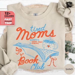 May include: Sand-colored sweatshirt with the text "tired Moms Book Club" in orange and blue. The design includes an open book, a coffee cup with a heart, and the phrase "Running on coffee, chaos & chapters."