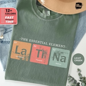 Op de afbeelding: Lichtgroen T-shirt met de tekst "-THE ESSENTIAL ELEMENT-" en de chemische symbolen La, Ti en Na in oranje, rode en groene vierkanten. Het shirt heeft ook de tekst "12+ COLORS" en "FAST PROCESSING TIME".