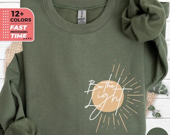 Op de afbeelding: Een groen sweatshirt met de tekst "Be the light" in witte letters en een zon-graphic in beige. De zon-graphic heeft lichtstralen die er vanaf komen.