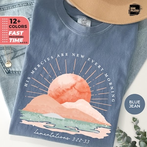Könnte beinhalten: Ein blaues Comfort Colors T-Shirt mit einem Aufdruck einer pfirsichfarbenen Sonne, die über Bergen aufgeht. Der Text "HIS MERCIES ARE NEW EVERY MORNING" und "Lamentations 3:22-33" sind auf dem Shirt aufgedruckt. Das Shirt wird auf einer blauen Jeans präsentiert.