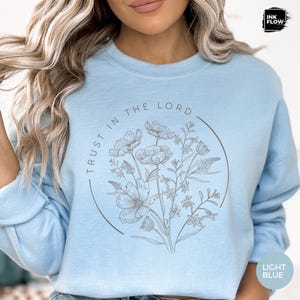 May include: Light blue sweatshirt with a floral design inside a circle. The text "TRUST IN THE LORD" is arched around the circle. The bottom right corner has the text "LIGHT BLUE".