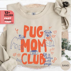 May include: A sand-colored crewneck sweatshirt featuring a graphic print with the orange text PUG MOM CLUB and the blue text life is better with pugs. The design includes two line-art pugs, a dog bowl, paw prints, hearts, and a bone icon.