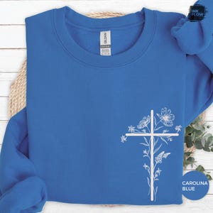 May include: A Carolina blue sweatshirt with a white floral cross design. The crewneck sweatshirt features a simple cross with delicate flower and leaf details. The text "Carolina Blue" is printed on the bottom right.