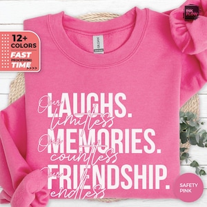 May include: A safety pink sweatshirt with the words "Our Laughs, Limitless Memories, Countless Friendship, Endless" in white script. The sweatshirt has a crew neck and long sleeves. A pink scrunchie is also visible.
