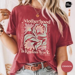 May include: Crimson t-shirt featuring the words "Motherhood Is Kindom Work" and floral graphics. The shirt includes text from Proverbs 31:25-27. The design incorporates a square floral arrangement in light pink and green.