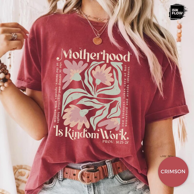 May include: Crimson t-shirt featuring the words "Motherhood Is Kindom Work" and floral graphics. The shirt includes text from Proverbs 31:25-27. The design incorporates a square floral arrangement in light pink and green.