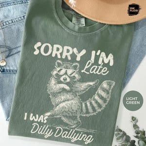 May include: Light green t-shirt with a raccoon graphic wearing sunglasses and the text "Sorry I'm Late, I Was Dilly Dallying" in white. The shirt is folded on a white surface with a hat and jeans.