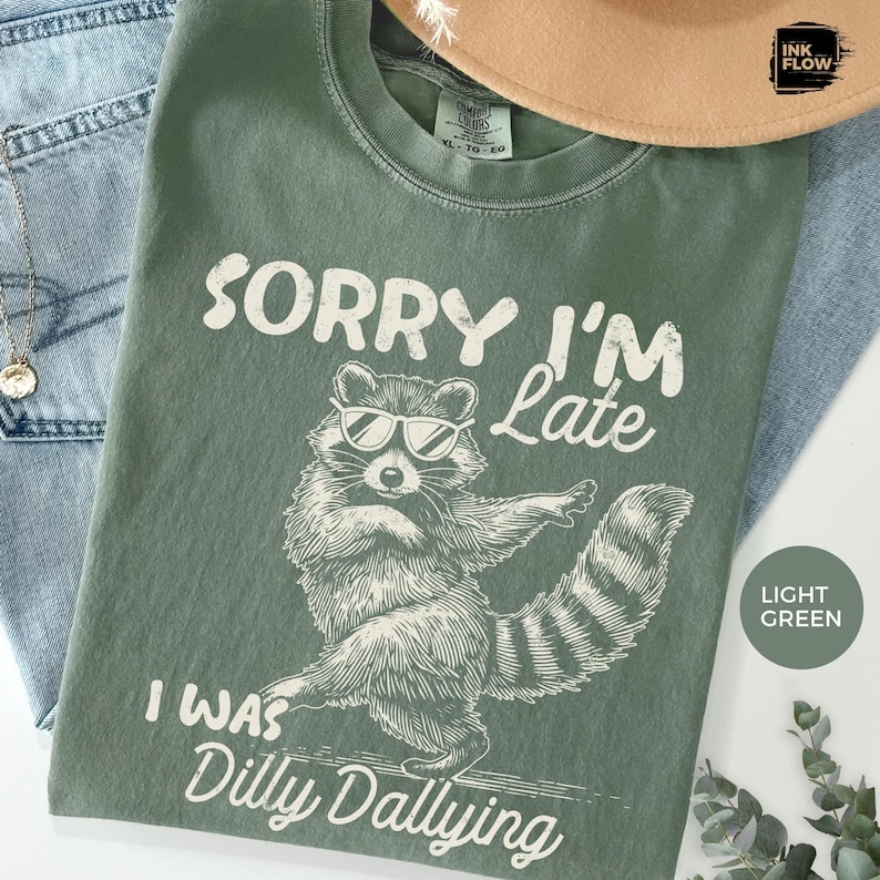 May include: Light green t-shirt with a raccoon graphic wearing sunglasses and the text "Sorry I'm Late, I Was Dilly Dallying" in white. The shirt is folded on a white surface with a hat and jeans.