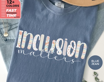 以下が含まれることがあります： 「Inclusion matters」の文字が装飾的なフォントでプリントされた、ブルーデニムカラーのTシャツ。「Inclusion」の文字は様々な模様と色で構成されています。Tシャツはクルーネックで半袖です。