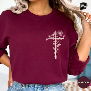May include: Maroon sweatshirt featuring a white floral cross design. The long-sleeved top has a crew neck and is made of a soft material. The word "MAROON" is printed on a circular label in the bottom right corner.