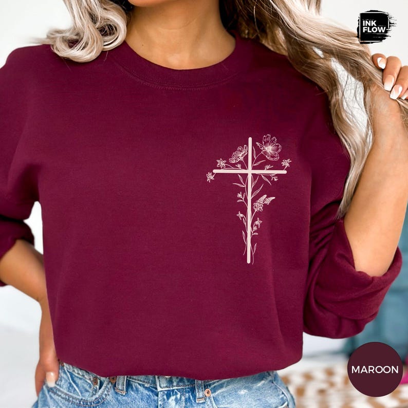 May include: Maroon sweatshirt featuring a white floral cross design. The long-sleeved top has a crew neck and is made of a soft material. The word "MAROON" is printed on a circular label in the bottom right corner.