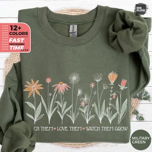 Op de afbeelding: Een groen sweatshirt met een witte afbeelding van bloemen en de tekst "CH THEM LOVE THEM WATCH THEM GROW". Het sweatshirt is verkrijgbaar in 12+ kleuren.