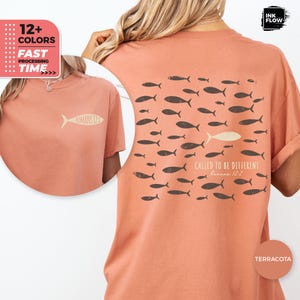 Puede incluir: Camiseta color terracota con gráficos de peces. La parte posterior muestra un grupo de peces oscuros con un pez claro y el texto "CALLED TO BE DIFFERENT Romans 12:2". La parte delantera tiene un gráfico de pez con el texto "ROMANS 12:2".