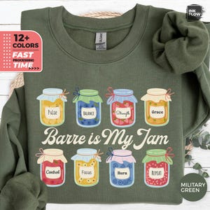 May include: Military green sweatshirt with the phrase "Barre is My Jam" and illustrations of eight jam jars, each labeled with words like "Pulse," "Balance," and "Strength." The sweatshirt also features text indicating multiple color options and fast processing time.