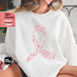 Könnte beinhalten: Weißes Sweatshirt mit einem rosafarbenen Banddesign, das aus kleineren rosafarbenen Bändern, Herzen und den Worten "Hoffnung", "Stärke", "Liebe" und "Kämpfen" besteht.