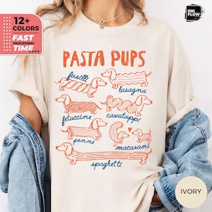 以下が含まれることがあります： オレンジ色のテキストで「PASTA PUPS」と書かれたグラフィックプリントが施されたアイボリーのTシャツ。デザインには、フジッリ、ラザニア、フェットチーネ、カヴァタッピ、ペンネ、マカロニ、スパゲッティなど、さまざまなパスタの形をした7匹のダックスフントが描かれています。