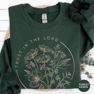 May include: Forest green sweatshirt with the text "TRUST IN THE LORD" in a circular design. The design includes a beige floral illustration. The sweatshirt is made of a soft material.