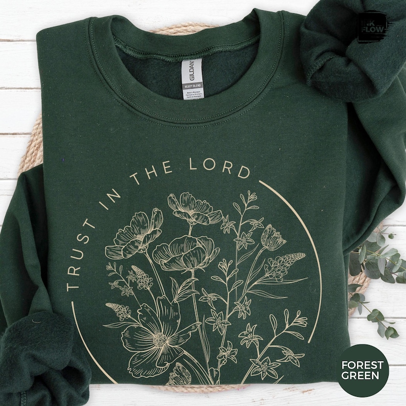 May include: Forest green sweatshirt with the text "TRUST IN THE LORD" in a circular design. The design includes a beige floral illustration. The sweatshirt is made of a soft material.