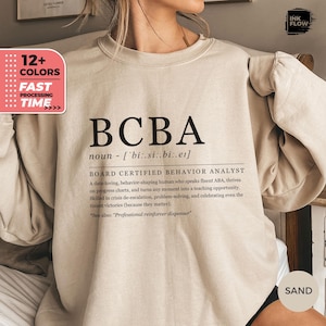 Könnte beinhalten: Ein beiger Pullover mit dem Text "BCBA" und der Definition "Board Certified Behavior Analyst". Der Text ist schwarz und die Definition ist in einer kleineren Schriftgröße.