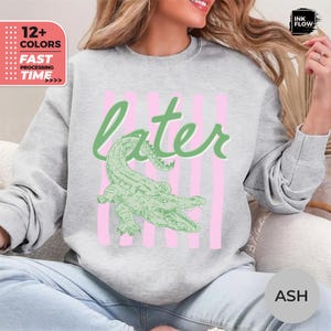 May include: A light grey crewneck sweatshirt featuring a graphic print of a green alligator positioned below the word later in a cursive green font. The design includes vertical light pink stripes behind the alligator and the text.
