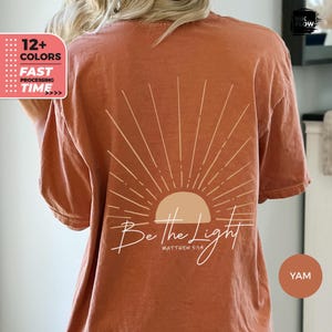 Könnte beinhalten: Ein rostfarbenes T-Shirt mit einem Sonnenstrahlen-Design und dem Text "Sei das Licht" mit dem Bibelvers "Matthäus 5,14".