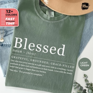 May include: A light green t-shirt with the word "Blessed" in white, and a definition. The shirt also features the text "Grateful, Grounded, Grace-Filled" and a quote. The shirt is displayed with a hat and jeans.