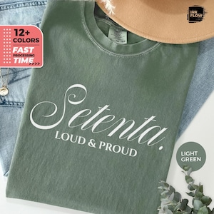 May include: Light green t-shirt with the word "Setenta" in white script, and the words "LOUD & PROUD" below. The shirt is folded on top of blue jeans. A tan hat and a necklace are also visible.