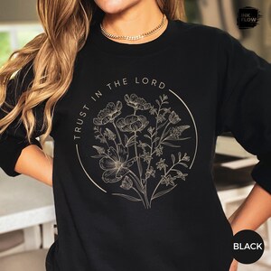 May include: Black sweatshirt with a floral design inside a circle. The words "TRUST IN THE LORD" are printed around the circle. The floral design is in a neutral color, and the sweatshirt is black.